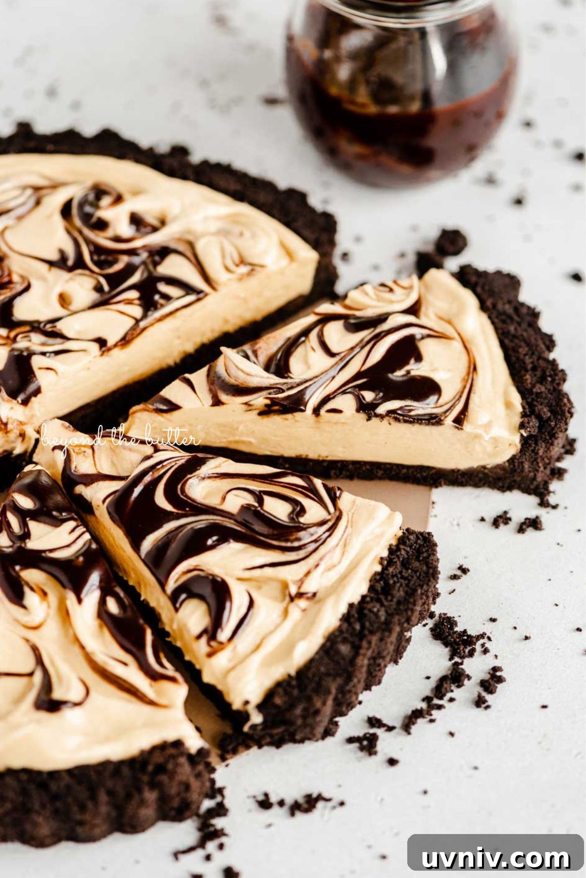 Round chocolate peanut butter tart swirled with chocolate syrup and cut into slices with a bowl of chocolate syrup next to it surround by a few chocolate sandwich cookie crumbs on a white speckled background.