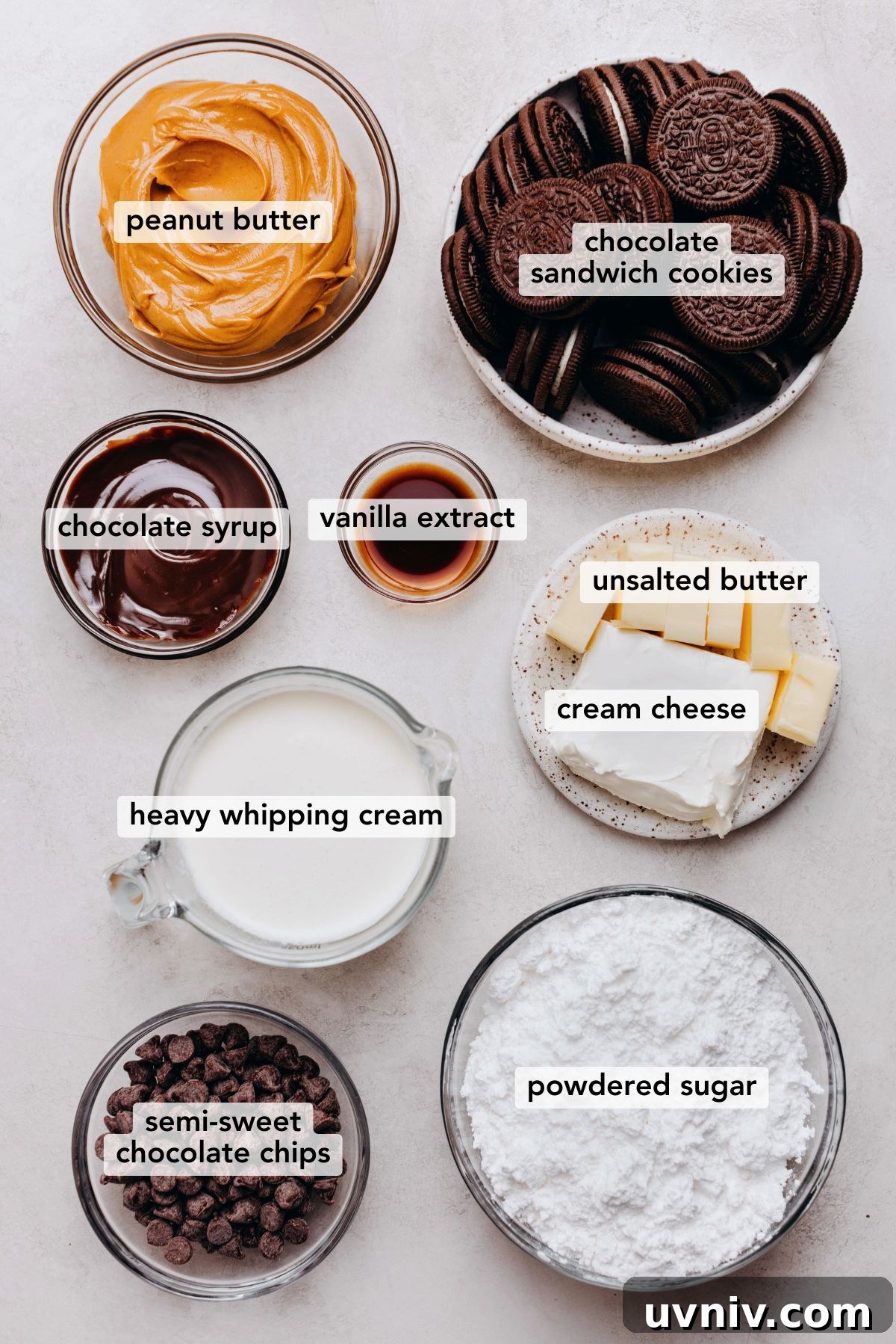 Ingredients in various size bowls labeled as peanut butter, chocolate sandwich cookies, chocolate syrup, vanilla extract, unsalted butter, cream cheese, heavy whipping cream, semi-sweet chocolate chips, and powdered sugar on a white marbled background.