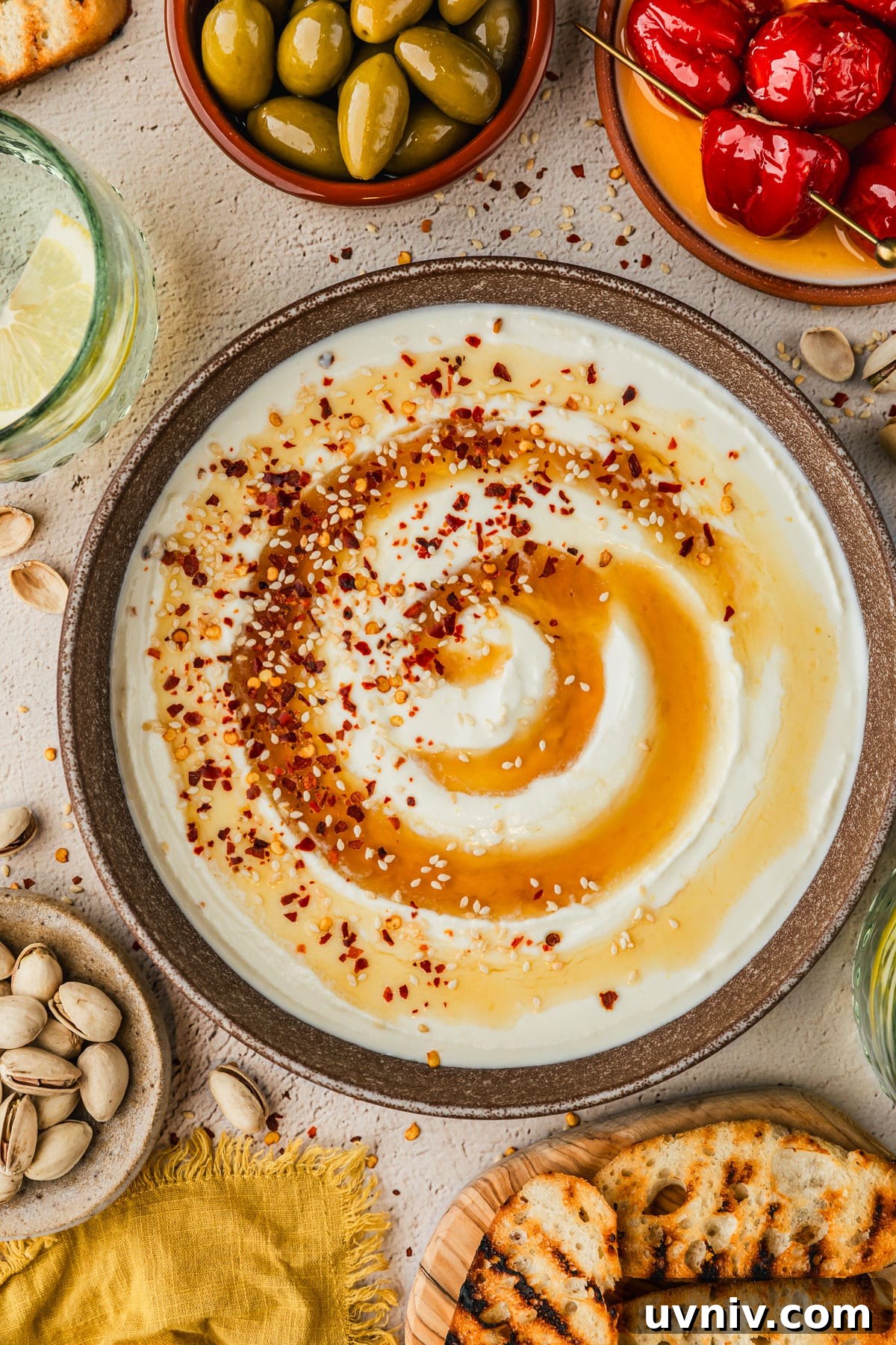 Whipped Feta with Honey in 5 Minutes — Creamy Dip Recipe 2 A brown bowl of whipped feta with honey on a tan counter next to glasses of a lemon cocktail, a wood board of bread, and brown bowls of pistachios, peppers, and olives.