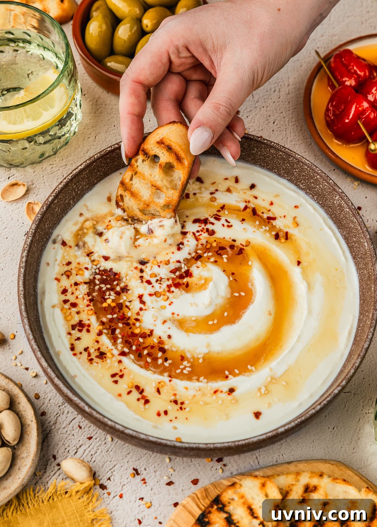 Whipped Feta with Honey in 5 Minutes — Creamy Dip Recipe 7 A hand dipping bread into a brown bowl of whipped feta with honey next to glasses of a lemon cocktail and brown bowls of olives, peppers, and pistachios with a tan background.