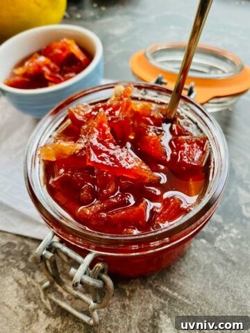 Close up of deep orange-red quince jam in a preserve jar with the lid opened and a teaspoon in