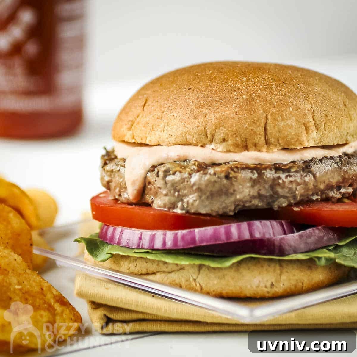 Creamy Sriracha Sauce Recipe for Burgers, Wraps & Sandwiches 8 Close up side shot of a burger with sriracha sauce on top on a clear plate with potato chips on the side.