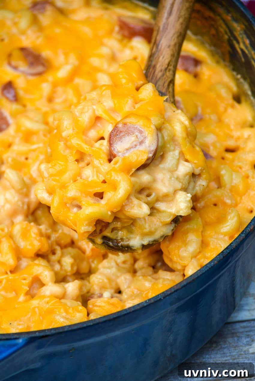 Firehouse-Style Macaroni and Cheese Recipe 6 a wooden spoon scooping firehouse mac and cheese with hot dogs out of a large blue pot