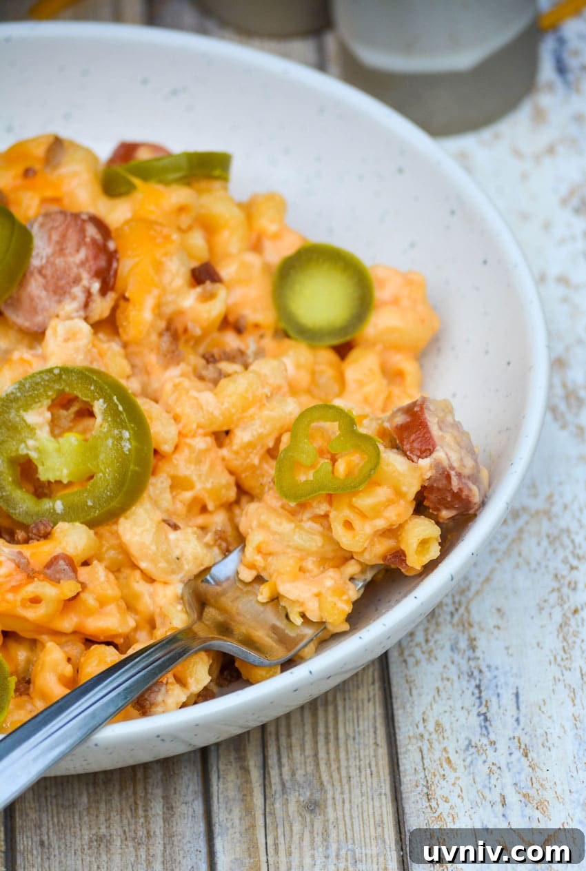 Firehouse-Style Macaroni and Cheese Recipe 7 a silver fork stuck in a bowl of baked macaroni and cheese with hot dogs topped with crumbled bacon and slices of jalapeno