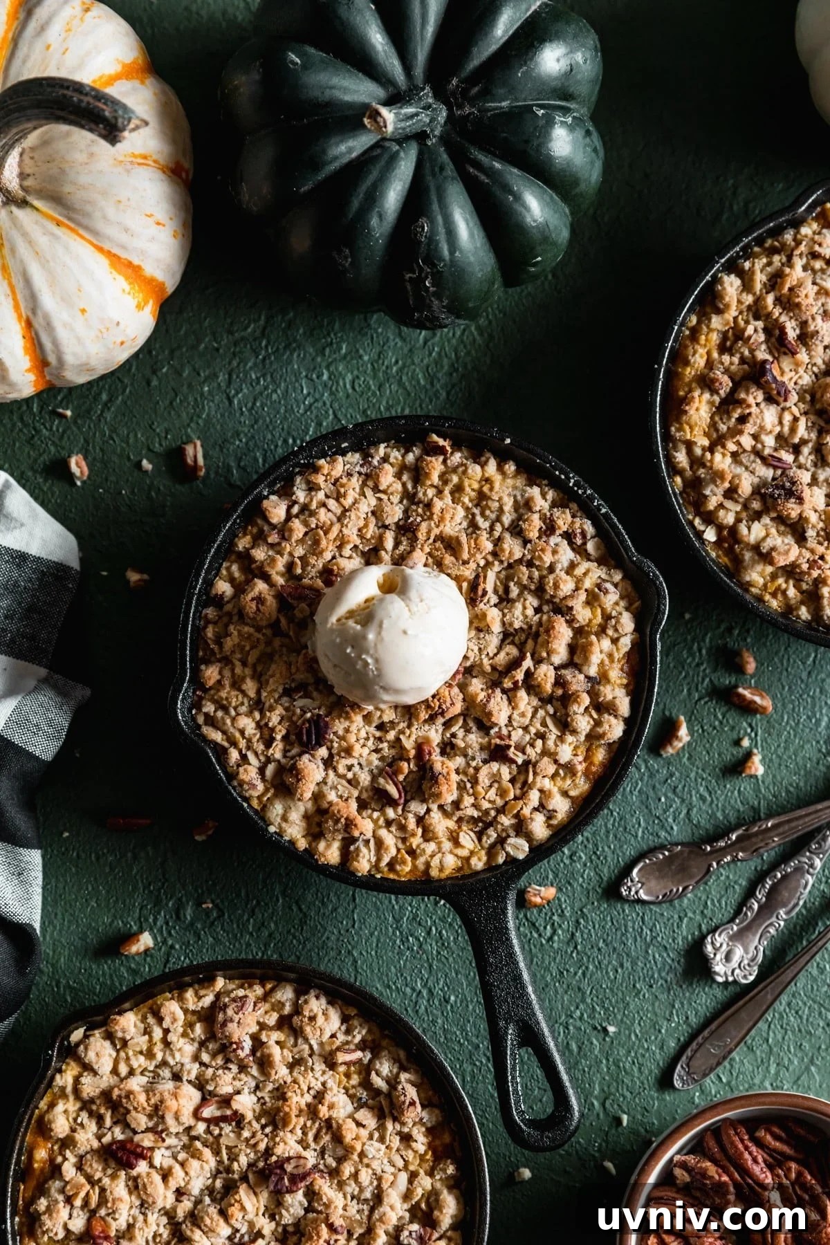An overhead image of a mini cast iron skillet with pumpkin crisp and a scoop of vanilla ice cream on a dark green table next to two more skillets, a black and white checkered linen, and vintage forks.