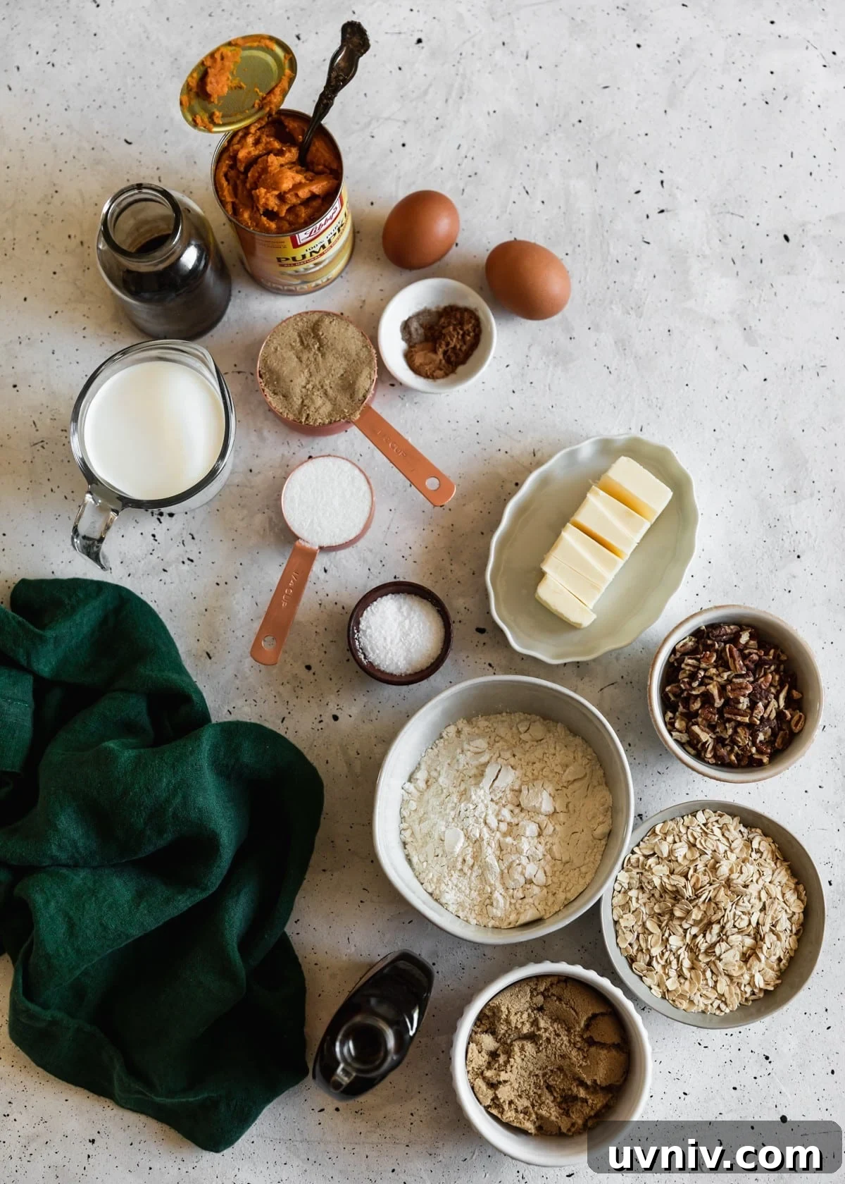 An overhead of various white and grey bowls with baking ingredients like flour, oats, pecans, sugar, and butter on a white speckled counter next to a can of pumpkin and dark green linen.