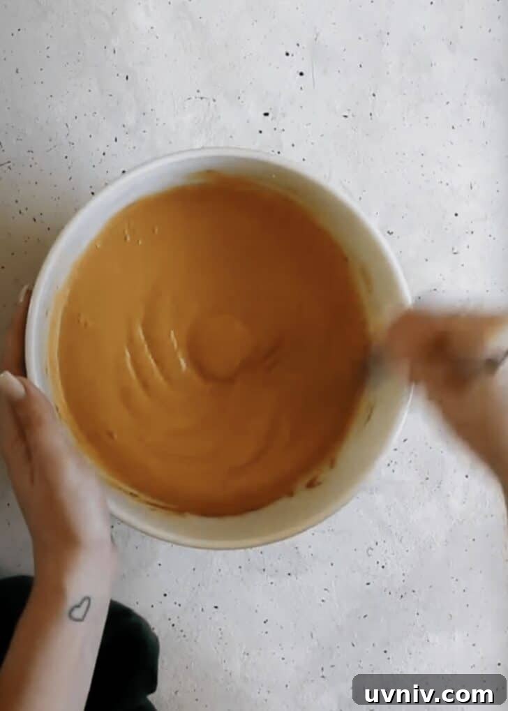 A hand using a whisk to mix pumpkin filling in a white bowl on a grey counter.