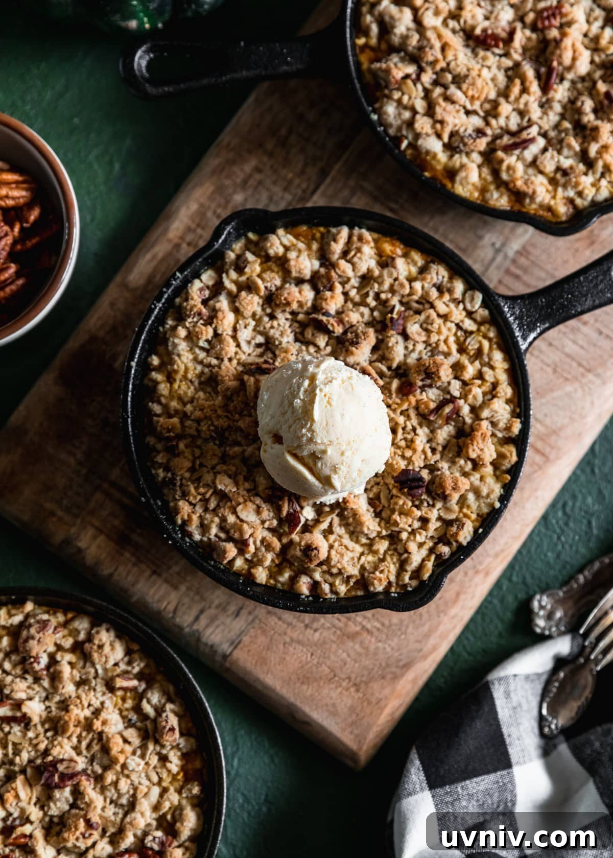 A closeup of a mini cast iron skillet with pumpkin crisp and a scoop of vanilla ice cream on a wood board next to another crisp. The board is on a green table next to a white bowl of pecans, a black and white linen, and vintage forks.