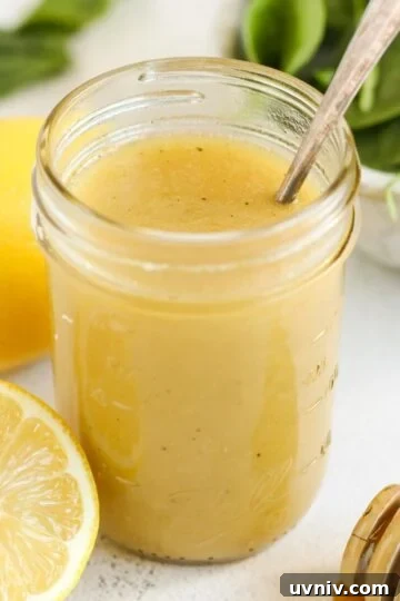 Lemon honey dressing in a mason jar with a sliced lemon, honey dipper and a bowl of spinach on the side.